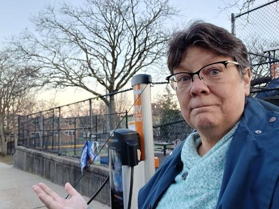 A white woman with brown hair and glasses, in a fleece shirt and a blue overcoat. I'm standing next to an EV charger that I'm using to charge my car. Behind me is the adjacent school playground, you can mostly see chain link fence and bare trees.