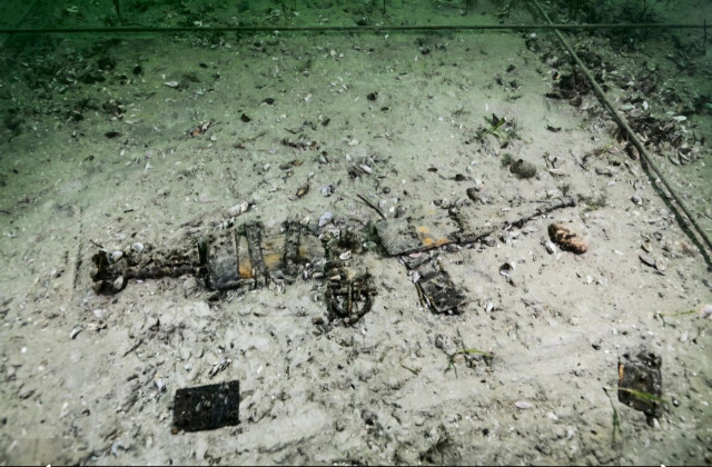 Underwater photograph of a remarkably preserved Roman sword, a so-called gladius, lying in its scabbard on a sandy seabed covered in green algae and sediment. Sand and small rocks surround it.