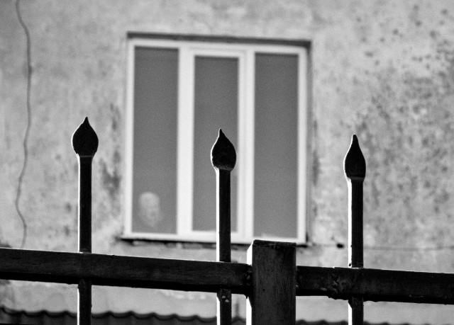 A black-and-white photograph of a metal fence in the foreground, its vertical bars topped with spear-like finials. Behind it, slightly out of focus, stands a weathered building facade with a three-pane window.

In the lower part of the window, an elderly woman is visible, her face soft and blurred as she looks out toward the street. The distance, the fence, and the shallow focus create a sense of separation, giving the scene a quiet, contemplative, slightly melancholic mood.