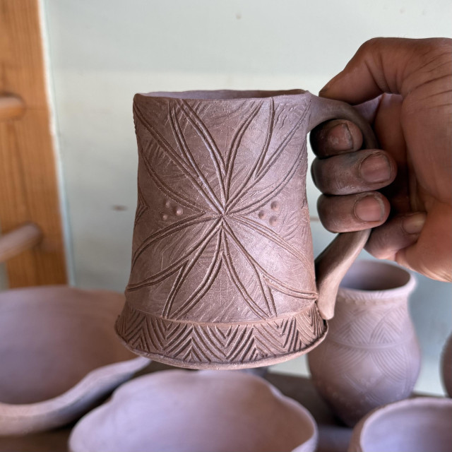 My hand holding a mug I’ve just carved a design into. It is brown and at the leather-hard stage of drying. The mug features various textures including a bold leaf motif, finely carved geometric patterns along the bottom and small raised dots. 