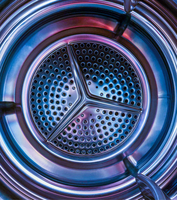 A straight-on close-up shot looking directly into the drum of a tumble dryer. The stainless steel interior glows with iridescent blue and purple hues. Three curved paddles divide the perforated back panel into equal sections, creating a strong three-way symmetry. Concentric rings of the drum surround the central disc. The lighting gives the mundane interior an almost otherworldly, sci-fi quality