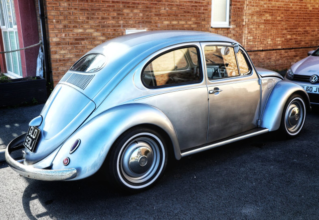 A well-preserved, silver Volkswagen Beetle parked on a tarmacked surface beside a brick building. The classic car features chrome hubcaps, whitewall tyres, and a vintage rear number plate reading 'GLD 931J'. Its iconic curved bodywork and split rear window highlight the model’s mid-20th-century design. The Beetle’s metallic silver paintwork gleams in the daylight, with another modern Volkswagen visible in the background.