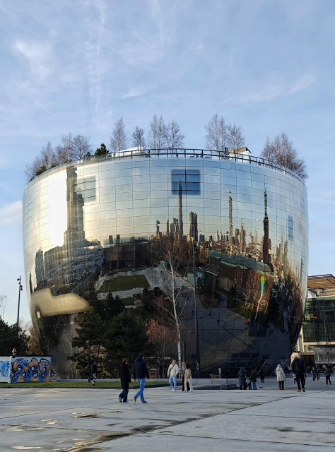 A photo of a large bowl-sgaped building with reflective glass panels and bare trees on the top. The glass reflects city buildings, construction cranes, people walking and a cloudy sky.