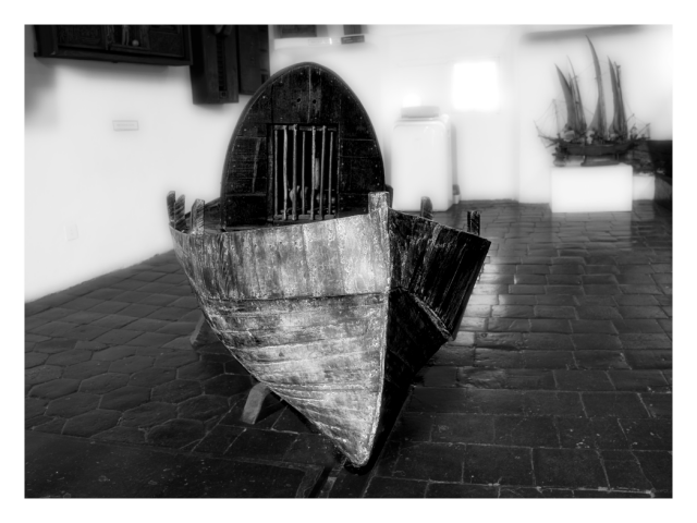 Indoor monochrome photography.
In the foreground, the bow of an unusual row-boat, moored on a cluster of terracotta tiles, faces the viewer, background exhibits various blurred artifacts from which one can guess a more ambitiously blooming sailing boat model.