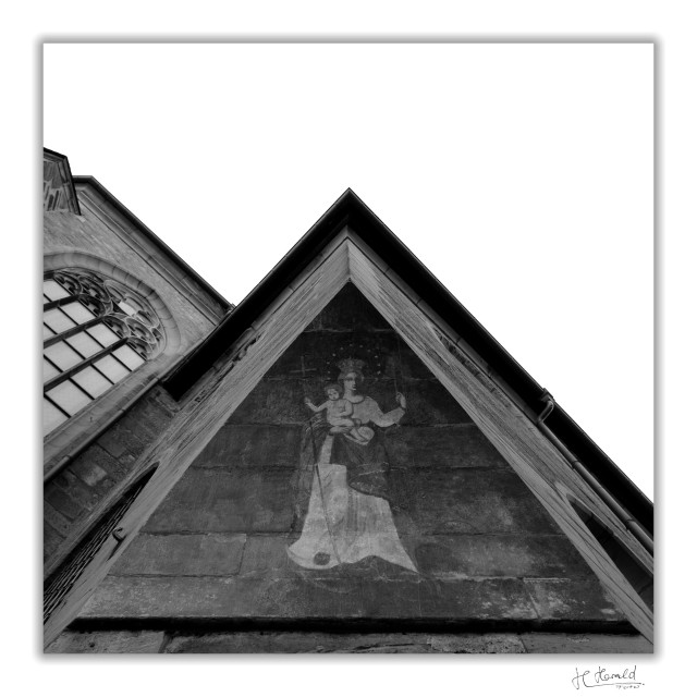 Square black-and-white image with a white frame of an outside corner of a church. The view looks upwards along the edge of the building. A painting of Mary and Jesus is depicted on a rounded overhang. To the left, you can see part of the church façade with a tall Gothic window.