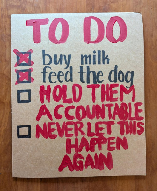 Handmade sign on a rectangle of cardboard, in black marker and red paint. “TO DO<br>☒ buy milk<br>☒ feed the dog<br>☐ hold them accountable<br>☐ never let this happen again”