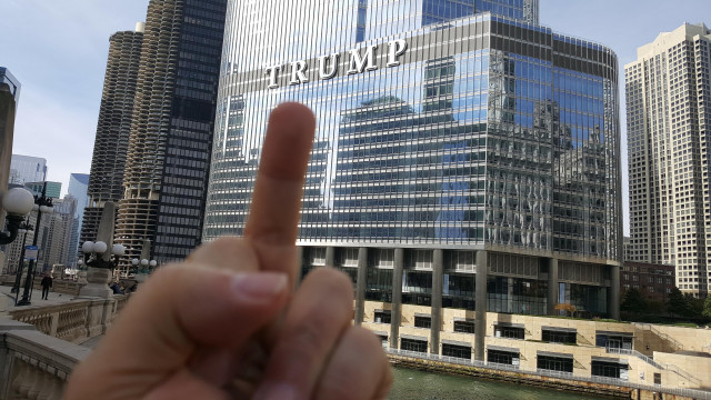 A hand in the foreground holds up a middle finger toward Trump Tower in downtown Chicago on a clear day.