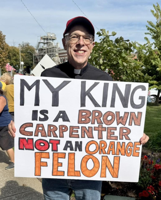 A smiling pastor wearing a black clerical shirt with a white collar and a red cap stands outdoors at a peaceful protest, holding a handmade sign that reads “MY KING IS A BROWN CARPENTER NOT AN ORANGE FELON” in bold black, red, and orange letters. The background shows a sunny day with trees, flowers, and a few people gathered, suggesting a public demonstration or rally. The message contrasts religious identity with political criticism, reflecting activism expressed through protest signage.