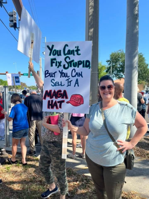 A smiling woman stands at a street protest during a “No Kings” demonstration, holding a handmade sign that reads, “You can’t fix stupid, but you can sell it a MAGA hat,” with the phrase “MAGA hat” illustrated in red. She wears sunglasses, a light green shirt, and a crossbody bag, standing near a traffic light pole while other protesters with signs gather behind her on a sunny day.