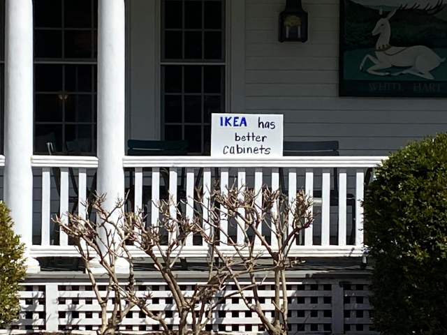 A handmade protest-style sign reading “IKEA has better cabinets” sits on the railing of a white wooden porch in front of a house. The sign, written in blue and black marker with “IKEA” emphasized, is placed between porch columns, with trimmed bushes and leafless branches in the foreground, giving the scene a quiet but pointed political tone.