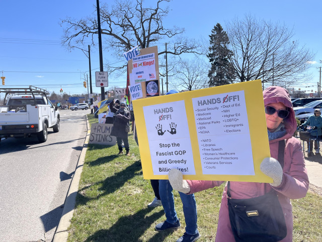 A woman wearing a winter coat with hood, mittens, and sunglasses is holding a protest sign reading "HANDS OFF! Stop the Fascist GOP and Greedy Billionaires' on the left side of the sign. And then it lists on the right side different areas to keep HANDS OFF! such as Social Security, Medicare, Medicaid, EPA, NOAA, Department of Education, NIH, Higher Education, etc., etc. 

It is a beautiful day and the sky is a bright blue. The trees are still bare. There are lots of people lining the street with their protest signs.