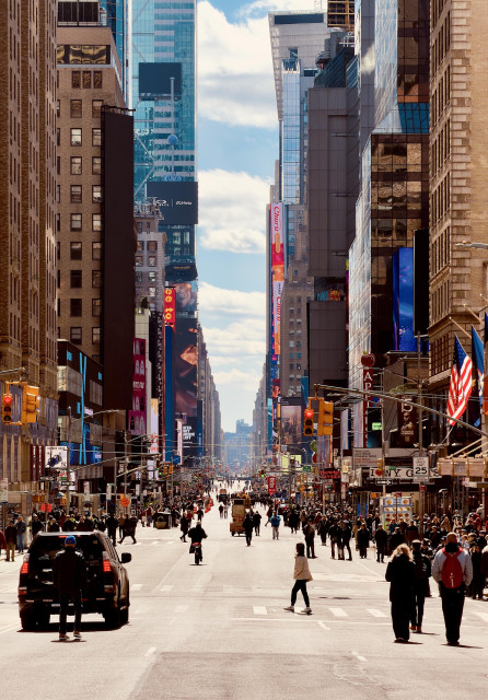 A color, wide-angle photograph in portrait orientation that captures a rare, expansive view looking south down Seventh Avenue from 52nd Street toward Times Square. The broad asphalt canyon is strikingly empty of vehicles, cleared for an upcoming protest march. In the foreground, a few scattered pedestrians and a cyclist cross the wide street, while a single black police SUV sits at a red light on the left. In the mid-ground, a dense crowd of people begins to swell, filling the street and sidewalks as they head south. The towering skyscrapers on either side create a deep linear perspective, drawing the eye toward the horizon where the blue sky and soft white clouds peek through the urban corridor. The architecture is a mix of classic stone facades and modern glass towers, many of which are adorned with the vibrant, glowing digital billboards characteristic of the Theater District. On the right, American flags flutter from a building near the "I LOVE NY GIFTS" shop and the Playwright Tavern. Bright yellow traffic lights hang over the empty lanes, their glow contrasting with the cool shadows cast by the buildings. The lighting is crisp and natural, with sunlight hitting the upper stories of the skyscrapers and casting long, soft shadows across the street, emphasizing the scale and temporary stillness of the normally bustling Manhattan thoroughfare.