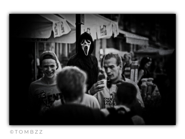A black-and-white street photograph with a dense, dark atmosphere, reminiscent of a frame from a horror movie. Heavy vignetting and deep shadows tightly frame the composition, heightening a sense of unease and entrapment.
​In the center of the frame is a group of young men. Although they occupy the central space, they are not in sharp focus—their silhouettes are soft, giving the scene a fleeting, almost dreamlike quality. A casual, joyful atmosphere prevails among them; smiles and animated discussion are visible. However, this image of ordinary human warmth is brutally disrupted by what is happening in the background.
​From behind, near a vertical pole, a dark figure emerges wearing the mask and costume from the movie "Scream." Due to the foreshortening of the perspective, the figure appears to be right behind the men, though it is actually at a distance. This motionless, eerie silhouette silently watches the group, introducing a theme of lurking evil and a threat of which the participants are entirely unaware.
​The foreground is heavily blurred, suggesting the perspective of someone spying on the scene from hiding. Other figures in the background are merely faint, blurry shapes, allowing all attention to focus on the key relationship: the contrast between the carefree smile of youth and the static, terrifying mask of death.
