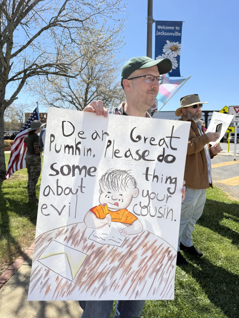 Photo of a white man in a ball cap holding a sign featuring the Peanuts character, Linus. He’s at a table writing a letter which says “Dear Great Pumkin [sic] , please do something about your evil cousin.”

In the background, other protesters are standing near the highway which runs past the town square. The sky is a beautiful, cloudless blue. 