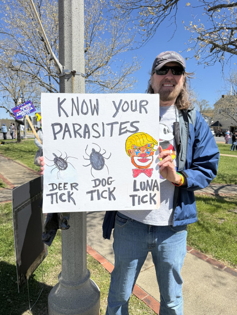 A man with long hair and a beard and wearing a ball cap holds a sign. “KNOW YOUR PARASITES” is at the top, and below are a small tick with “deer tick” below it, a larger tick with “dog tick” below it, and a clown with orange hair and a bow tie with “Luna Tick” beneath it. The beautiful blue sky and a budding tree are in the background.