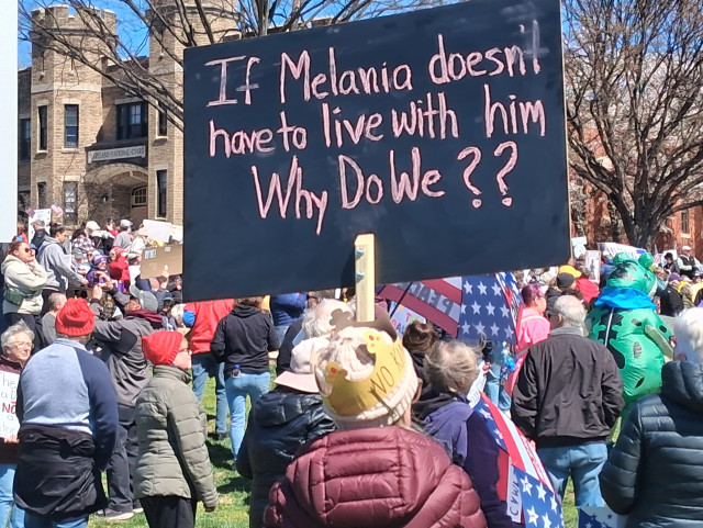 A sign that says "If Melania doesn't have to live with him Why De We??" Plenty of people surrounding it.