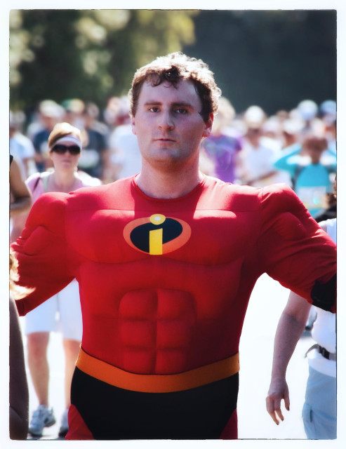 A picture taken in 2009 at the annual Bay to Breakers footrace in San Francisco. It shows a man in a red Mr. Incredible muscle suit, leading a pack of runners through Golden Gate Park. As I hold up my camera to take his picture, he counters my move with arms outstretched, ready to hug or perhaps crush me.
