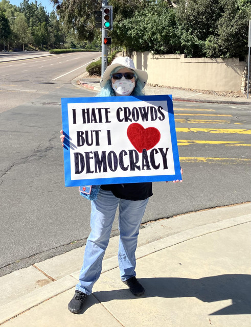 White woman with blue hair, N95 mask and a protest sign that reads “I hate crowds but I ❤️ democracy” standing on a street corner 