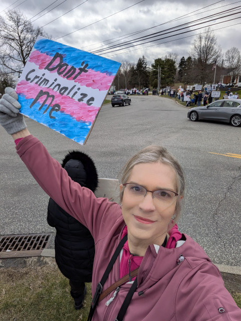 A selfie of a white trans woman holding a sign at the March 28, 2026 No Kings protest. The sign is hand painted with the words "Don't criminalize me" written over a stylized trans flag.