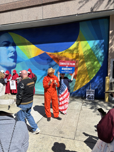 Man dressed in an orange prison jumpsuit. He’s handcuffed and wearing a Trump wig. His face is painted orange. Next to him a woman holds a sign saying “Convicted felon, or democracy, that’s the choice.” 