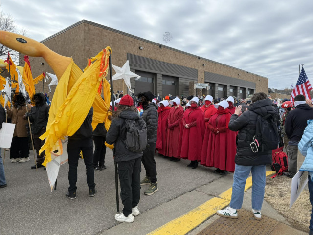 Photo of protesters, one group in red robes and white headpieces of The Handmaid’s Tale, a giant yellow bird puppet in front of them