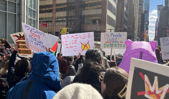 Within a protest march down 7th Avenue of NYC on a cold, sunny afternoon. Signs: “No thrones, No crowns, No kings”; “You can’t bomb your way out of the Epstein files”; “No kings since 1776”; “The people united will never be defeated”