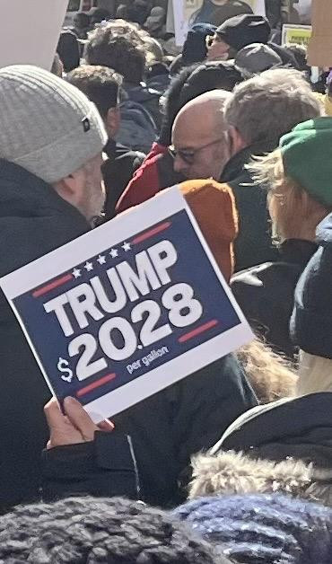 Within a protest march down 7th Avenue of NYC on a cold, sunny afternoon. Sign: A “Trump 2028” campaign poster, modified to read “Trump $20.28 per gallon”