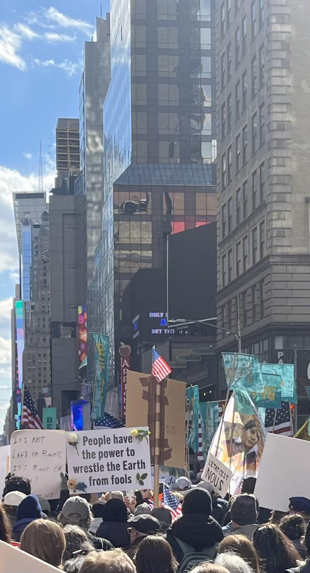 Within a protest march down 7th Avenue of NYC on a cold, sunny afternoon. Signs: “People have the power to wrestle the Earth from fools”; “It’s not left or right, it’s right or wrong”; “L’état c’est nous”