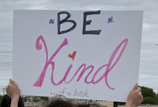 A hand holds up a large sign with the words "Be Kind ... of a bitch" written in bold, colorful letters. The background appears cloudy, and some people are partially visible.