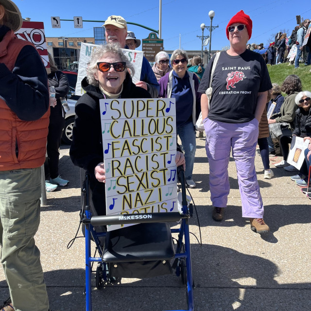An older woman smiles, holding a sign that reads "Super callous fascist racist sexist Nazi Potus."