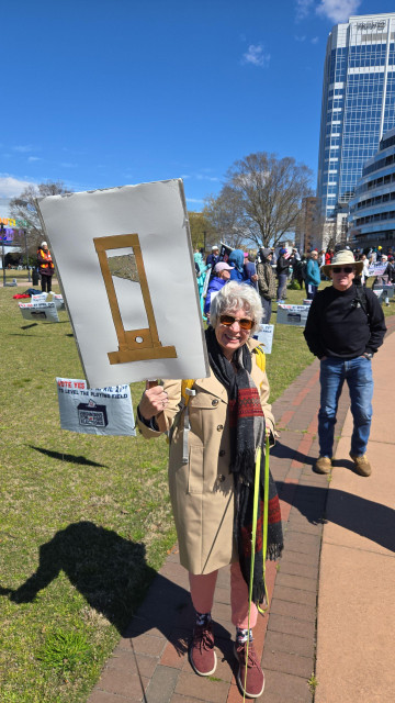 a 'no kings' protester, a smiling, white-haired, grandmother, holding a sign that is just a picture of a guillotine.