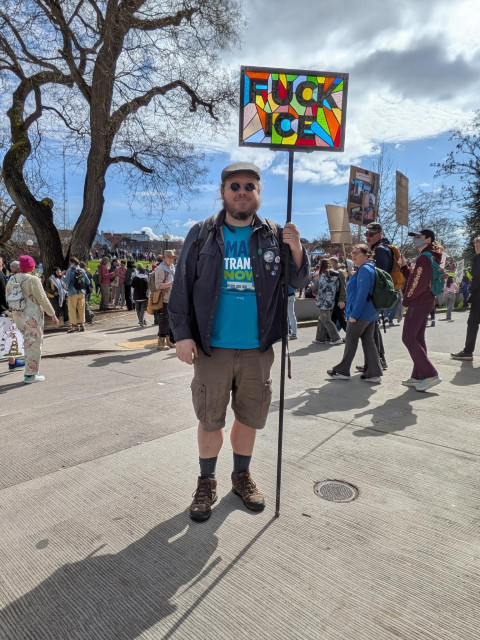 A scruffy looking guy in shorts, holding a 12" x 18" stained glass "FUCK ICE" sign that is mounted in a 3/4" steel frame, which has a removable 6' pipe bolted to it for use in a protest context. He is standing on the edge of a park, with hundreds of protesters behind him.