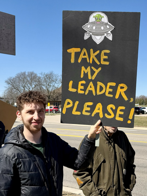 A No Kings protester with a sign with a UFO saying, “Take My Leader, Please!”