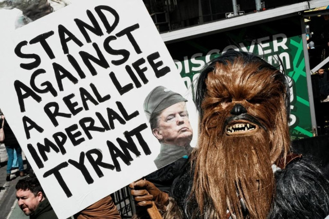 The person in the costume is dressed as Chewbacca from Star Wars, but the sign they are holding features Donald Trump edited to look like a Galactic Imperial Officer (wearing the distinct grey uniform and cap of the Galactic Empire).

The use of Chewbacca is deliberate here; in the Star Wars lore, the Wookiees were enslaved and oppressed by the Empire, making him a symbol of the "Rebellion" or resistance against an "Imperial Tyrant."

Imperial Tyrant Sign
STAND AGAINST A REAL LIFE IMPERIAL TYRANT