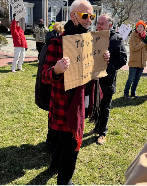 Elderly man with a thin mustache holding a cardboard sign