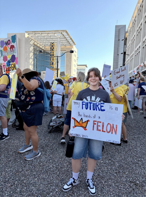 This is a photo of my 15 year old granddaughter at the No Kings protest in Las Vegas, Nevada. She's holding the sign she made that says - My Future is in the hands of a felon. There's a picture of a crown with an X through it. In the background are dozens of people participating with different signs and costumes. 