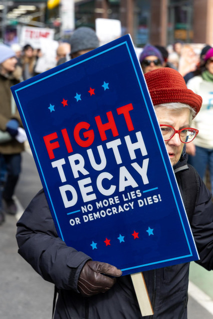 A white-haired woman in a red hat carrying a blue protest sign with the words ‘Fight Truth Decay’ printed on it in red and white.
