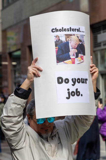 A man carries a sign with a photograph of Donald Trump eating a hamburger and the words “Cholesterol … do your job” written on it.
