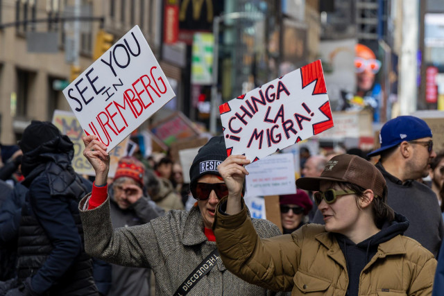 A crowd of protesters. Two people in front hold signs that read “See you in Nuremberg” and “¡Chinga la migra!”

