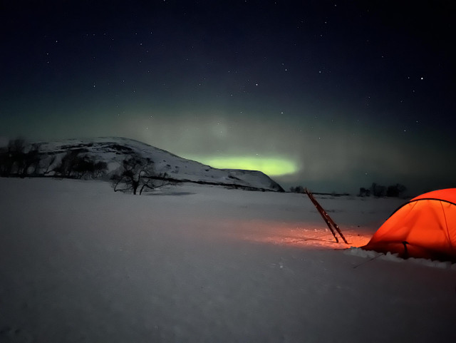 A red illuminated tent, at the horizon there’s a snow covered mountain and green northern lights behind it. There’s some stars visible in the sky. 