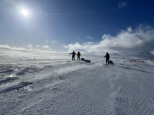 And icy, frosty landscape with three people on skis. There’s some snow drift visible close to the ground. 