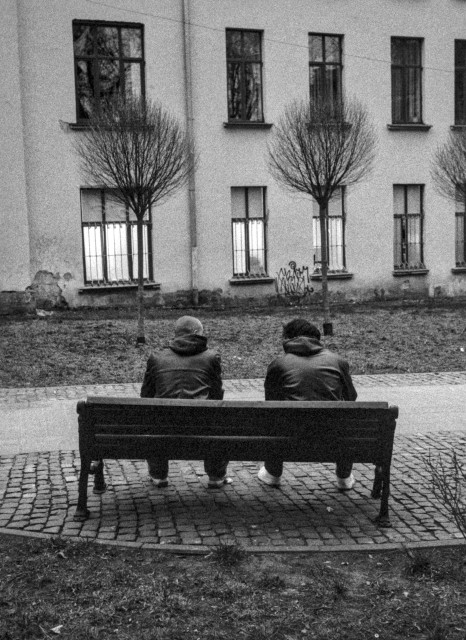 A grainy black-and-white photograph shows two men sitting side by side on a wooden bench, seen from behind. They both wear dark jackets and slightly hunched postures, facing a quiet courtyard. In front of them stands a row of small, leafless trees, evenly spaced along a narrow strip of ground. Behind the trees rises a plain building facade with tall rectangular windows, some covered with bars. The cobblestone pavement in the foreground adds texture, while the overall mood feels subdued and contemplative, as if the two figures are sharing a silent moment together.