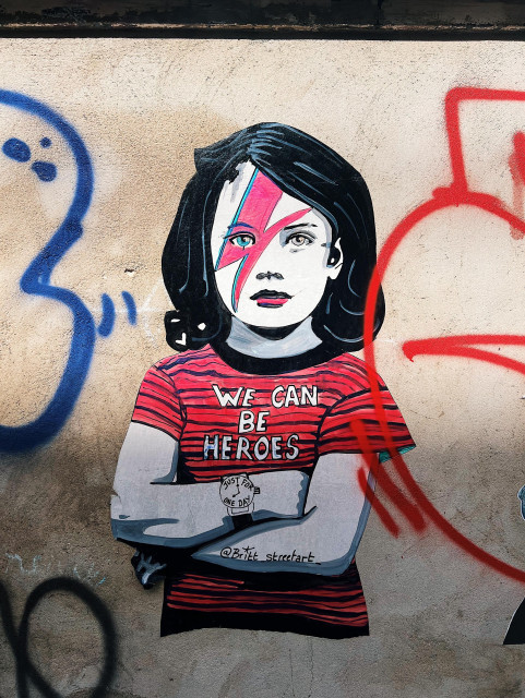 A photo of a graffiti on a wall showing a young girl with Bowie / Ziggy Stardust inspired makeup; she’s wearing a red & black striped t-shirt that reads “we can be heroes” and on her watch are the words: “just for one day” and on her other arm there’s the credit : @_britt_streetart_