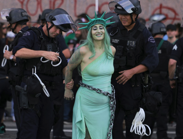 Photo of a woman dressed as the Statue of Liberty,  in a light green dress with matching hair, makeup, and crown to imitate the aged copper patina of the statue.  She wears a chain around her waste.  Two police officers, larger than her, are in the process of zip-tying her hands behind her back.  The police wear black uniforms with side-arms and riot helmets.  More police are seen in the background.  Liberty lady has her eyes closed and a smile of resignation, holding her head high. 