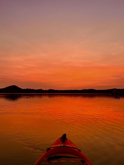From the point of view of sitting in a kayak. You see the tip of an orange kayak pointed into a deep red sunset that permeates the sky and is reflected off the water. The water is mostly still, but has ripples that highlight the deflections giving some depth to the photo. Taken at dusk so there is only a few more minutes of light yet to paddle back.