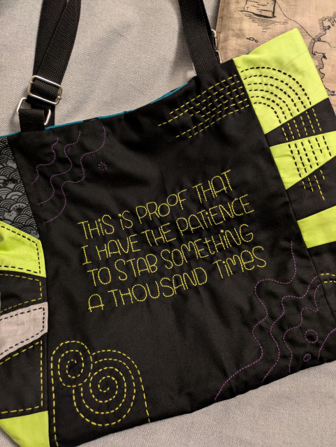 Black fabric shoulder bag with a patchwork design featuring green lime and patterned panels, decorated with colorful stitched embroidery. The front has yellow embroidered text reading: “THIS IS PROOF THAT I HAVE THE PATIENCE TO STAB SOMETHING A THOUSAND TIMES,” surrounded by decorative stitched swirls and lines.