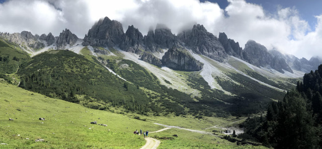 A panorama of great tooth like craggy mountain peaks rising from a grassy green valley, forested hills and dusty highs. Clouds are rising behind the peaks. In the centre bottom, a path, where tiny people can be spotted afar.
