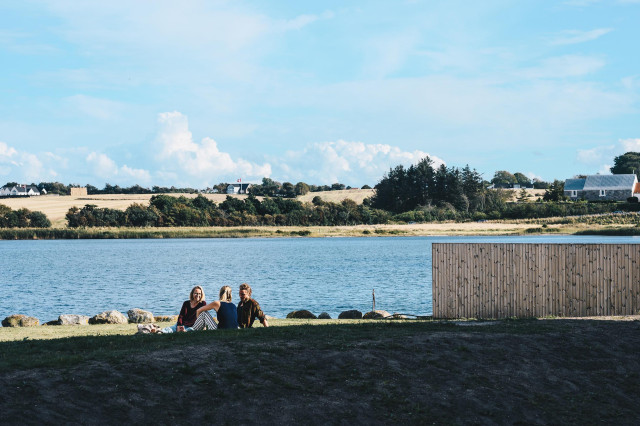 Tre mennesker sidder på græsset ved vandkanten på en sommerdag.
