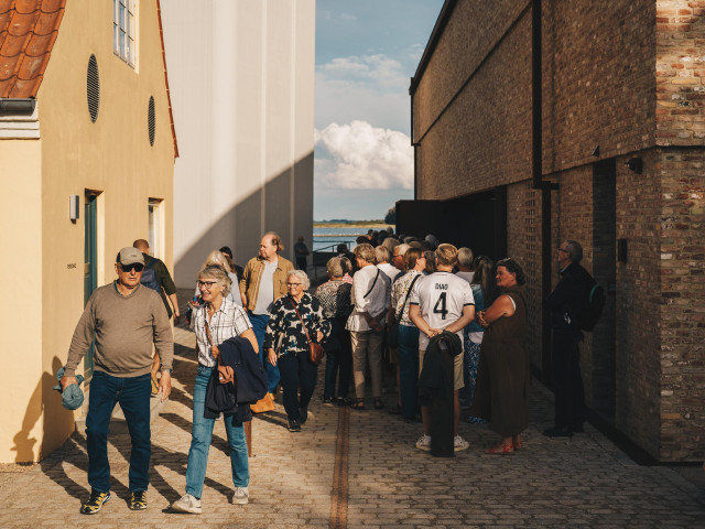 En gruppe mennesker går imellem gule bygninger på en sommerdag.