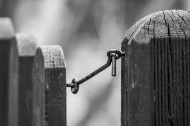 Schwarz weiß Aufnahme eines Gartentürchens mit Haken, der geschlossen ist. Das Türchen plus Pfeiler sind aus Holz, der Fokus liegt auf dem Riegel.